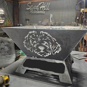 Puede incluir: Un gran brasero de metal oscuro con un diseño de cara de perro recortada. El brasero tiene forma cuadrada con lados angulados y una base robusta. El logotipo de Sand Hill Metalworks es visible en el fondo.