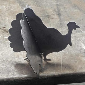 Könnte beinhalten: Metall-Truthahn-Silhouette, aus dunklem Metall geschnitten. Der Truthahn steht mit ausgebreiteten Schwanzfedern. Das Metall hat eine dunkle, matte Oberfläche. Der Truthahn ist ein dekorativer Gegenstand.