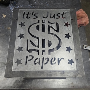 Puede incluir: Un letrero metálico con la frase "It's Just Paper" recortada, con un signo de dólar y estrellas. El letrero está hecho de metal gris y probablemente sea para exhibición en interiores o exteriores.