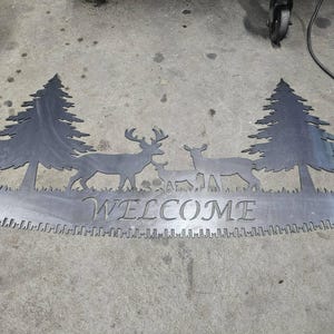 May include: A metal welcome sign featuring a deer family and two pine trees. The word "WELCOME" is cut out in the center, above a saw blade design. The sign is made of grey metal.