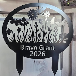 May include: Black metal garden stake featuring a floral and butterfly design within an oval frame. The text "Bravo Grant 2026" is displayed below the floral design. The stake has two pointed prongs for ground insertion.