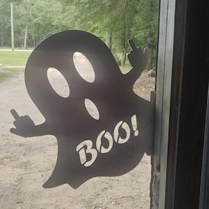 Könnte beinhalten: Ein Metall-Geist-Ausschnitt mit dem Wort "BOO!" und einer Hand, die den Mittelfinger zeigt. Der Geist hat drei ovale Augenausschnitte und ist an einem Holzpfosten befestigt. Der Hintergrund zeigt Bäume und einen Feldweg.