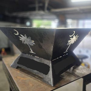 May include: A black metal fire pit with a square, angled design. The sides feature cut-out designs of a crescent moon and a palmetto tree. The fire pit is set on a metal table in a workshop setting.