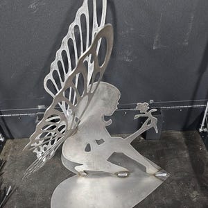 May include: A silver metal sculpture of a fairy with butterfly wings. The fairy is seated on a heart-shaped base and holding a flower. The wings have an open design. The sculpture is made of silver-coloured metal.