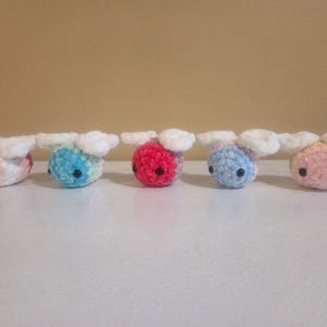 May include: Five small, round, crocheted plush toys shaped like bees, each featuring a soft, fuzzy texture, two white wings, and two black bead eyes. The bees are arranged in a row, displaying various colors including peach, blue, pink, and white.