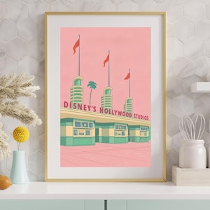 Op de afbeelding: Een ingelijste kunstdruk met Disney's Hollywood Studios. Het kunstwerk heeft een retro-stijl met een roze lucht, groene en blauwgroene gebouwaccenten en rode vlaggen. De tekst "DISNEY'S HOLLYWOOD STUDIOS" staat bovenaan het gebouw.