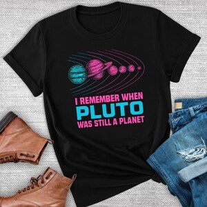 May include: Black t-shirt featuring a colorful graphic of planets and the text "I REMEMBER WHEN PLUTO WAS STILL A PLANET". The design includes a teal planet, a pink planet with rings, and other pink planets. The text is in pink and teal.