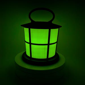 May include: A black metal lantern with a green glow emanating from the inside. The lantern is sitting on a white base.