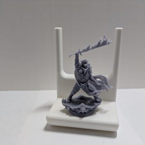 May include: A gray 3D printed miniature of a figure holding a flaming sword. The figure is wearing armor and a cloak. The miniature is standing on a circular base.