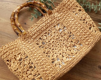 May include: A woven straw tote bag with a bamboo handle. The bag features a detailed openwork design in a natural tan color. The handle is made of curved bamboo, adding a rustic touch. The bag is rectangular and appears to be handmade.