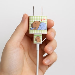 May include: A white phone charger with a green gingham pattern and a cartoon capybara design. The capybara is sleeping under a blue blanket with hearts above it. The charger has a small capybara illustration on the cord.
