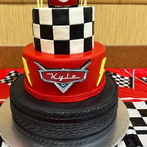 Op de afbeelding: Een drielaagse rode, zwarte en witte taart versierd met een ruitpatroon, een rode auto en een bliksem. De middelste laag heeft de naam "Kyle" op een rood auto-embleem. De onderste laag is versierd om eruit te zien als een band.