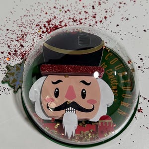 Può includere: Un ornamento a forma di sfera in plastica trasparente con un  interno di un  cavatappi rosso e oro glitterato. La sfera è circondata da glitter rosso e oro. Il testo "Countdown to Christmas" è scritto sulla sfera.