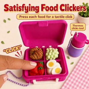 May include: A bright pink plastic lunchbox container with four compartments containing miniature food items: a croissant, green grapes, strawberries, and sliced hard-boiled eggs. A separate purple and pink thermos is attached by a chain.