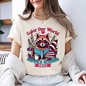 May include: A white t-shirt with a graphic of a raccoon wearing glasses and reading a book. The graphic also includes the text "Color Our World" with a heart, "Summer Reading 2025", and other colorful details.
