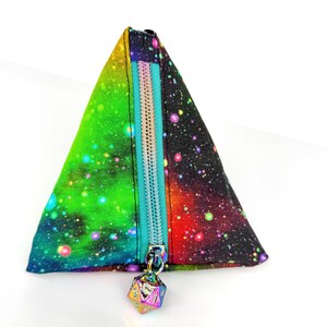 May include: A triangular pouch with a galaxy print in shades of green, blue, red, and purple. It features a teal zipper and a multi-colored die charm. The pouch is designed to stand upright.