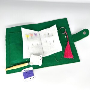 May include: A green leatherette sewing kit, open to show a white felt needle holder with needles and butterfly-shaped pins. Includes a pink tassel, a wooden needle threader, and white and purple thread spools.
