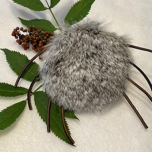 Puede incluir: Un pompón de piel gris y blanco con correas de cuero marrón. El pompón está unido a un cordón de cuero marrón.