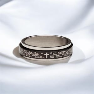 May include: A black and silver spinning ring with the Lord's Prayer engraved around the band. The ring features a cross design.