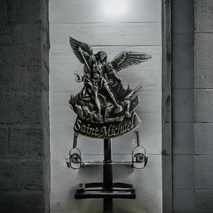 Könnte beinhalten: Eine silberne Statue des Heiligen Michael, der einen Drachen tötet, montiert auf einem schwarzen Metallständer mit zwei silbernen Wandleuchten. Die Statue steht vor einer weißen Wand mit der Inschrift "Saint Michael" auf dem Sockel.