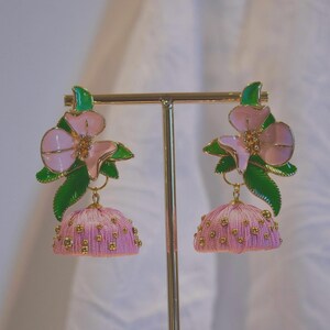 May include: A pair of dangle earrings featuring a pink flower with green leaves at the top, connected to a pink dome-shaped jhumka base decorated with small gold-colored beads. The earrings are displayed on a gold-toned metal stand.
