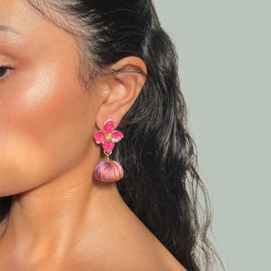 May include: A close-up of a pink floral earring with a gold-colored post. The earring has a flower-shaped top and a bell-shaped bottom with pink and gold threads. The earring is worn on a person's ear.