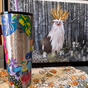 May include: A stainless steel travel tumbler with a colorful floral design, a black lid, and a purple straw. The tumbler is next to a framed illustration of a forest scene with a mythical creature.
