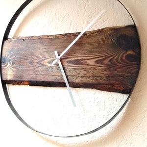 Könnte beinhalten: Eine rustikale Holzuhr mit einem Metallringrahmen und weißen Zeigern. Das Holz hat eine dunkelbraune Farbe und eine natürliche Holzmaserung.