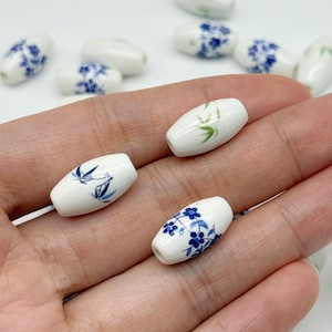 May include: A collection of white barrel-shaped beads featuring delicate hand-painted designs in blue and green. The patterns include small blue floral clusters and slender green bamboo stalks. Each bead has a smooth, glossy finish and a central hole.