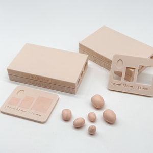 Könnte beinhalten: Ein Set aus hellbeigen Silikonformen und kleinen ovalen Tonstücken. Die rechteckigen Formen haben Aussparungen mit der Beschriftung 10mm, 12mm und 15mm. An der Seite der Formen steht der Text 10-12-15mm zur Kennzeichnung der Hohlräume.