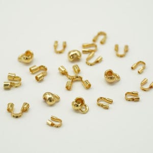 May include: A collection of small, gold-colored jewelry findings, including clasps, connectors, and decorative beads. The components are scattered on a white surface, showcasing various shapes and designs for jewelry making.