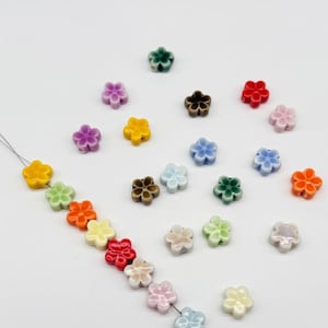 Puede incluir: Una colección de pequeñas cuentas multicolores con forma de flor. Algunas cuentas están ensartadas en un hilo fino, mientras que otras están esparcidas. Las cuentas presentan varios colores pastel y vibrantes, incluyendo amarillo, verde, naranja, rojo, morado, azul y blanco.