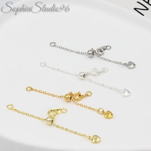May include: Close-up of four adjustable necklace extenders in silver and gold tones. Each extender features a small heart charm and a ball bead. The extenders are on a white surface.