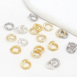 May include: A collection of gold and silver-colored jewelry findings, including open jump rings and clasps. The findings are scattered on a white surface, with some pieces overlapping. The jewelry components are small and shiny, reflecting light.