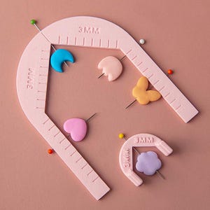 以下が含まれることがあります： 3MMの目盛りが付いたライトピンクのプラスチック製縫製ガイド2点。大きいガイドはU字型で、小さいガイドはコンパクトなアーチ型です。どちらも青い月、ピンクのハート、オレンジの蝶などのカラフルな粘土の形が縫い針で貫かれています。