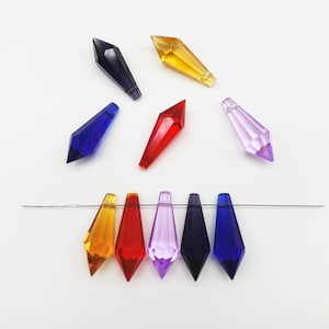 Puede incluir: Una colección de cuentas facetadas en forma de punta, similares a cristales, en varios colores que incluyen amarillo, rojo, azul, morado y azul marino oscuro. Cinco de estas cuentas están dispuestas en una fila horizontal sobre un hilo metálico plateado.