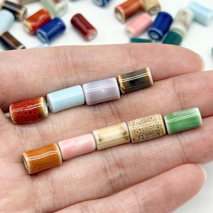 Könnte beinhalten: Eine Sammlung kleiner, zylindrischer Keramikperlen in verschiedenen Farben und Oberflächen, darunter gesprenkeltes Rot, helles Blau, Lavendel, Braun, Orange, Rosa, Creme und Grün, mit einem Haufen weiterer loser Perlen im Hintergrund.