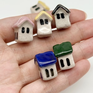 May include: A collection of five miniature ceramic house figurines, each featuring a white base with black painted windows and doors, topped with roofs in various colors including pink, yellow, black, blue, and green.