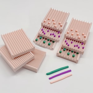 Peut inclure: Un ensemble de plateaux rectangulaires rose clair avec des rainures parallèles, certains marqués 3.5mm et 4mm. Les plateaux contiennent de petites perles sphériques pêche, violettes et vertes. Trois tiges cylindriques en pâte sont posées à côté.