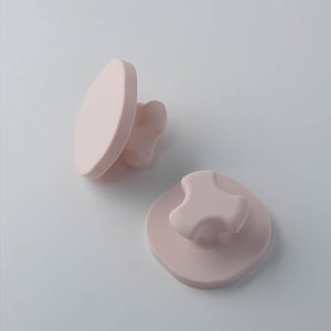 May include: Two pale pink knobs with a smooth, matte finish. One knob is shown from a side angle, revealing its circular face and three-pronged base, while the other is positioned flat, displaying its rounded top and three-lobed grip design.
