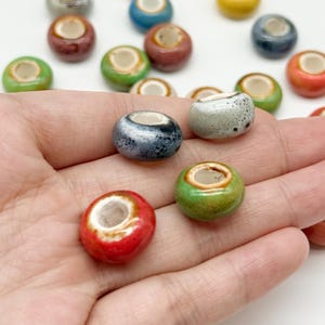 May include: A collection of small, round, ceramic beads in various glossy colours including red, green, blue, and speckled grey. Each bead features a smooth, rounded exterior with a central hole, showcasing a glazed finish and a white inner rim.
