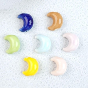 Puede incluir: Una colección de siete cuentas con forma de media luna en varios colores, incluyendo azul oscuro, bronceado, verde claro, azul claro, rosa pálido, amarillo brillante y melocotón suave. Cada cuenta presenta un acabado liso y brillante y un diseño curvo.