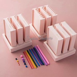 Puede incluir: Un conjunto de tres soportes rectangulares de color rosa claro con partes superiores ranuradas etiquetadas con medidas de 2mm a 4,5mm. Junto a los soportes hay varias varillas de arcilla delgadas y multicolores y pequeñas cuentas esféricas de arcilla.