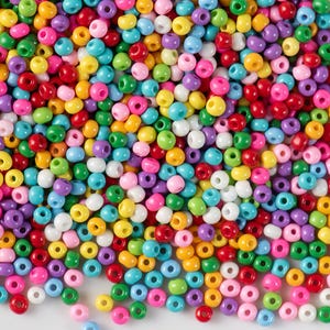 Könnte beinhalten: Nahaufnahme einer Ansammlung kleiner, runder Perlen in verschiedenen Farben, darunter Rot, Gelb, Grün, Blau, Lila, Rosa und Weiß. Diese Perlen werden wahrscheinlich für Bastelarbeiten und Schmuckherstellung verwendet. Die Perlen sind verstreut.