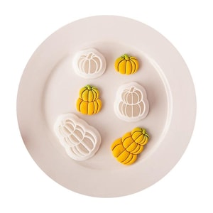 Può includere: Una collezione di tagliabiscotti in plastica bianca a forma di zucca e i corrispondenti biscotti gialli a forma di zucca. Il set include una forma di zucca singola e una forma di zucca doppia impilata, disposte su un piatto chiaro.
