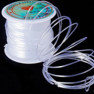 May include: A spool of clear, stretchy beading cord. The spool has a white base and a teal and red label with Japanese text. The cord is wound around the spool and also trails off to the side.