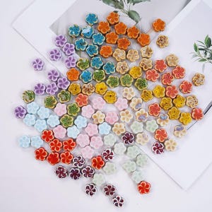Könnte beinhalten: Eine große Sammlung kleiner, fünfblättriger blumenförmiger Perlen in einer Vielzahl von Farben, darunter Orange, Blau, Grün, Gelb, Rosa, Rot, Violett und Weiß. Jede Perle hat eine glänzende Oberfläche und ein zentrales Loch zum Auffädeln.
