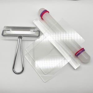 May include: A set of clear acrylic crafting tools including a metal-handled roller, a cylindrical rolling pin with purple and pink rings at each end, and two square acrylic sheets of different sizes for flattening and smoothing materials.