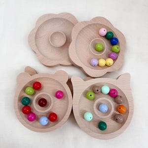 Könnte beinhalten: Vier helle Holzschalen mit erhöhtem Rand und kreisförmigen Vertiefungen in der Mitte. Zwei Schalen sind blumenförmig, zwei haben die Form von Tierköpfen mit Ohren. Jede Schale enthält kleine, bunte, glatte, kugelförmige Perlen.