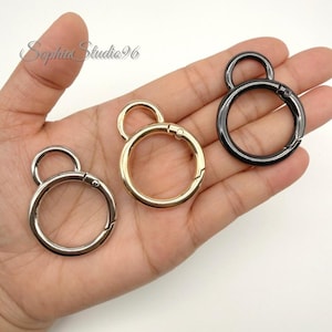May include: Three circular spring-loaded metal clasps in silver, gold, and black finishes. Each clasp features a large main ring with a smaller attached loop for attachment. The metal rings have a smooth, polished surface and a functional spring gate.